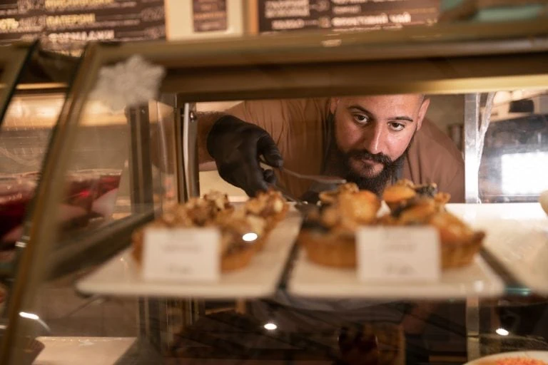 Bakery owner reaching into bakery display fridge and getting a pastry to serve to a customer